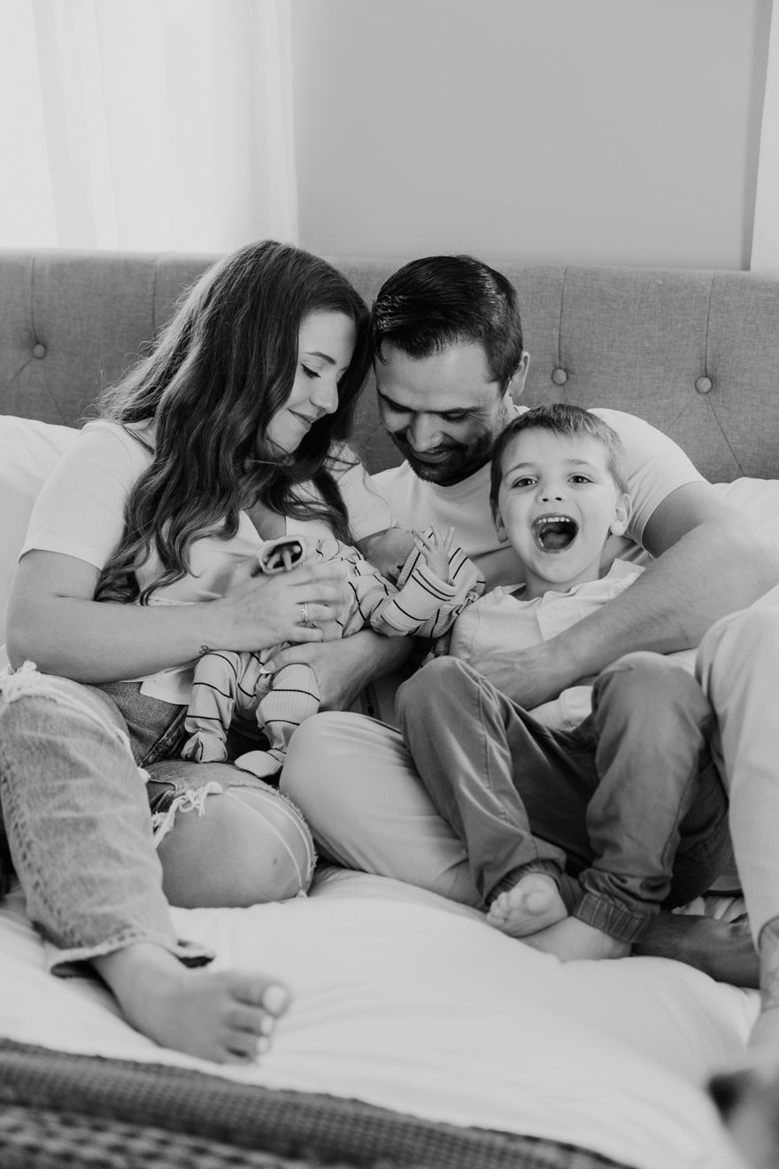 Black and white photo of a happy family of four sitting on a bed, parents laughing with toddler and baby.