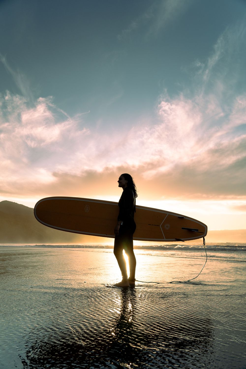 portrait of a woman standing in front of the sun with surfboard under her arm looking up at the ocean in Raglan, New Zealand Aotearoa