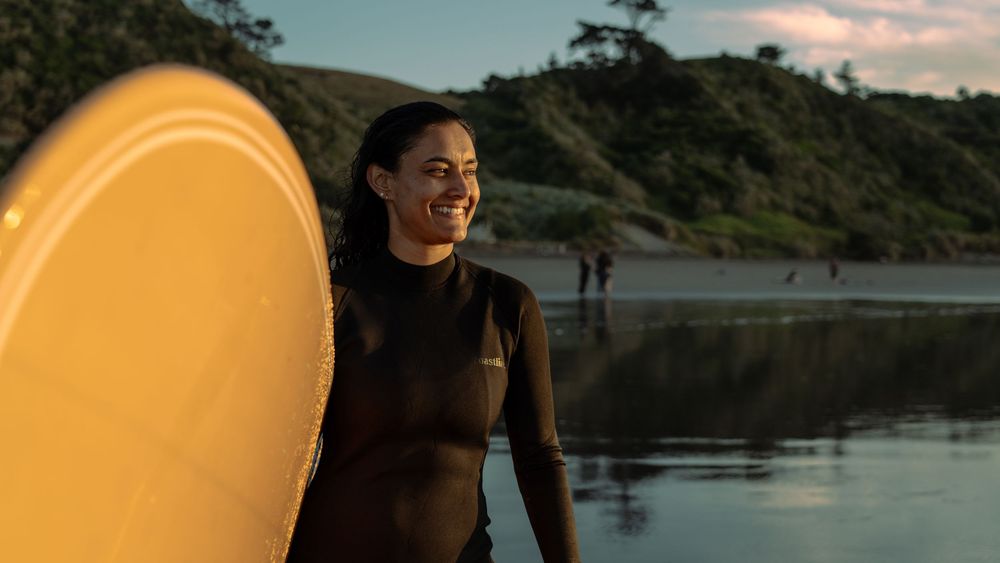 portrait of a woman standing smiling at the ocean with surfboard under her arm in Raglan, New Zealand Aotearoa