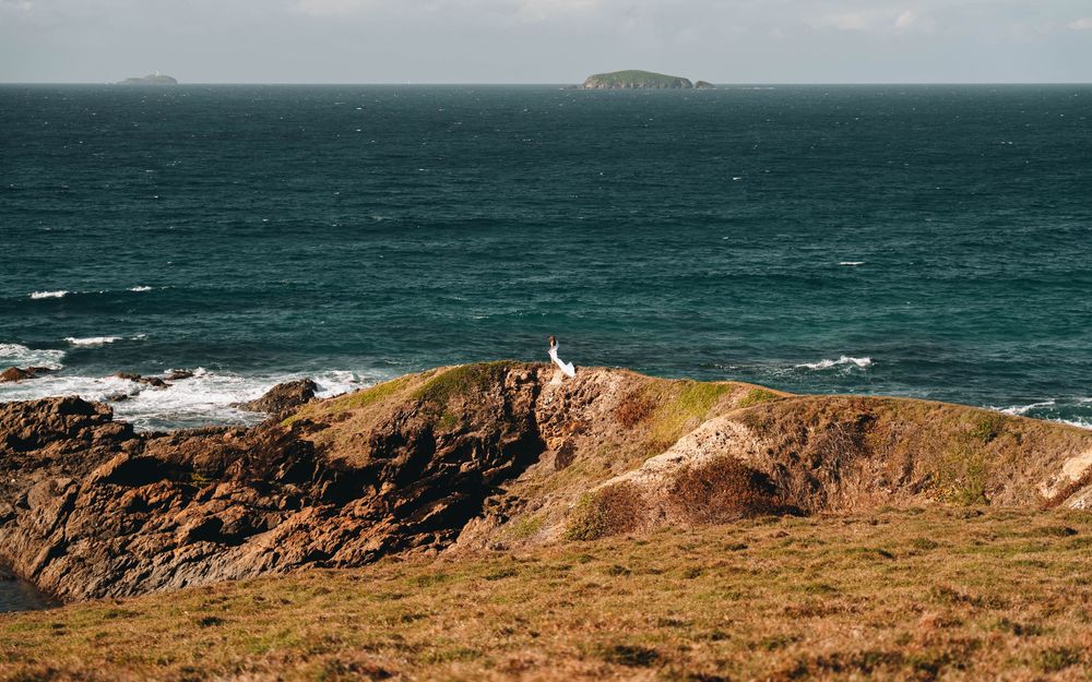 woman in long white dress standing far away on a cliff with the blue rugged coastline of NSW and the waves coming onto shore