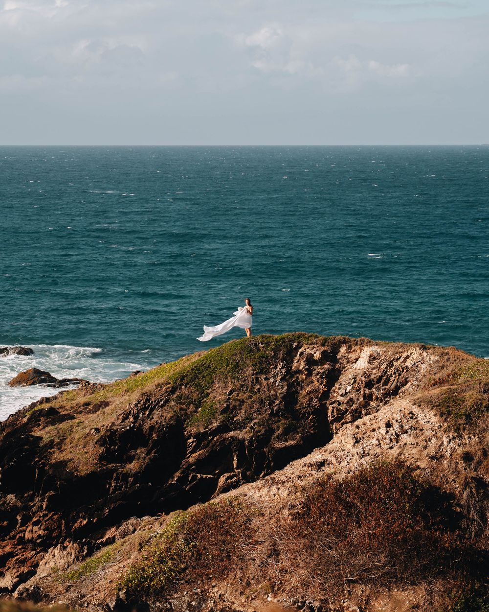 woman in long white dress standing far away on a cliff with the blue rugged coastline of NSW and the ocean behind her