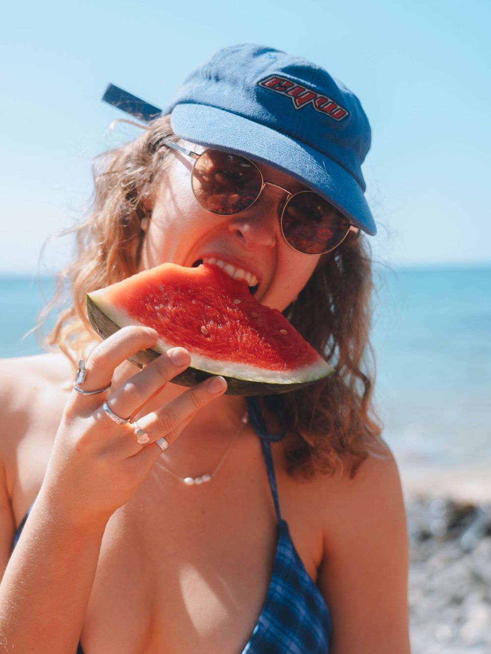 a girl in blue matching bikini and cap eating watermelon showing rings on her fingers in the NSW summer at the beach with ocean in the back