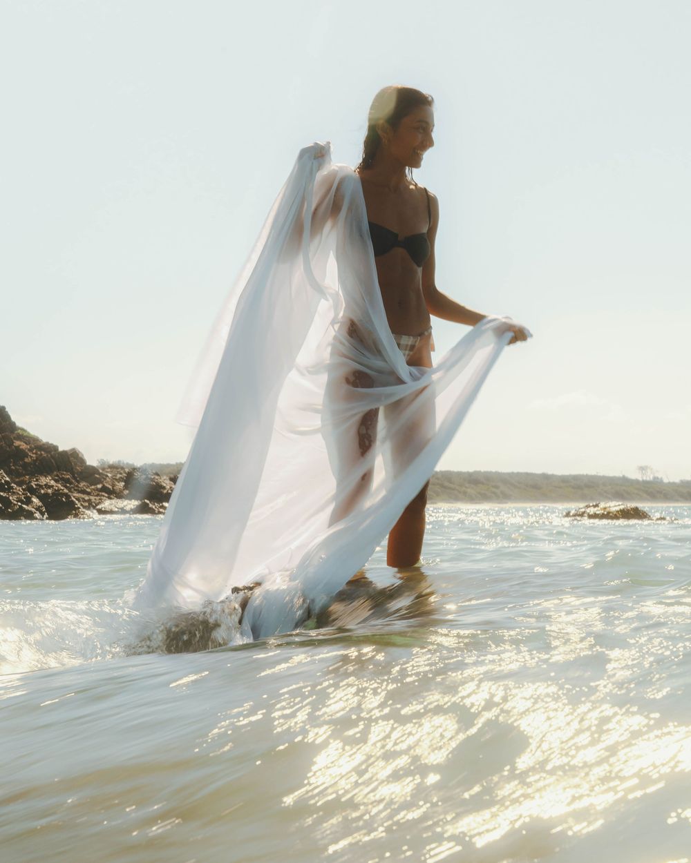 Girl holding a satin sheet along her body standing on a rock in the ocean with a wave making the wave look milky at Mid North Coast NSW