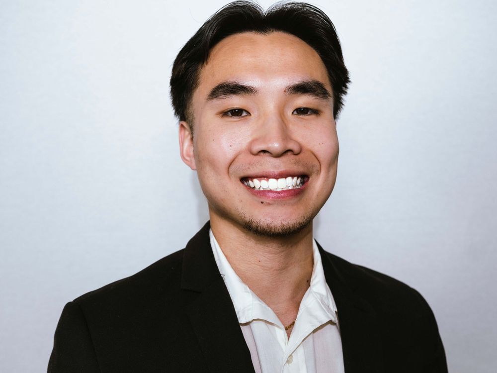 Young professional man in black blazer smiling confidently against a light gray background in a headshot photo.