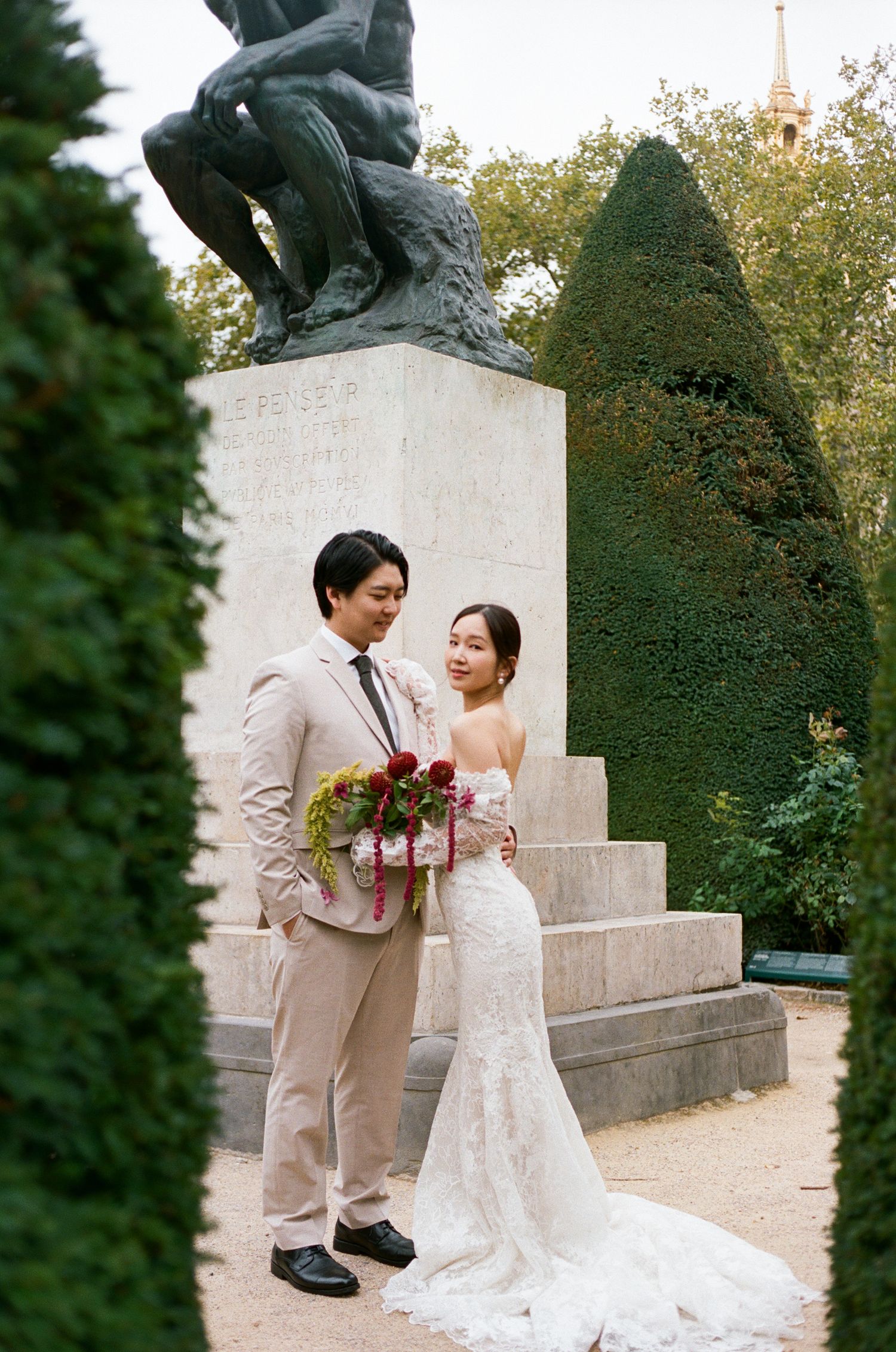 Asian couple in wedding attire poses before a bronze statue in musee rodin. Paris asian wedding. Paris editorial wedding photography, film
