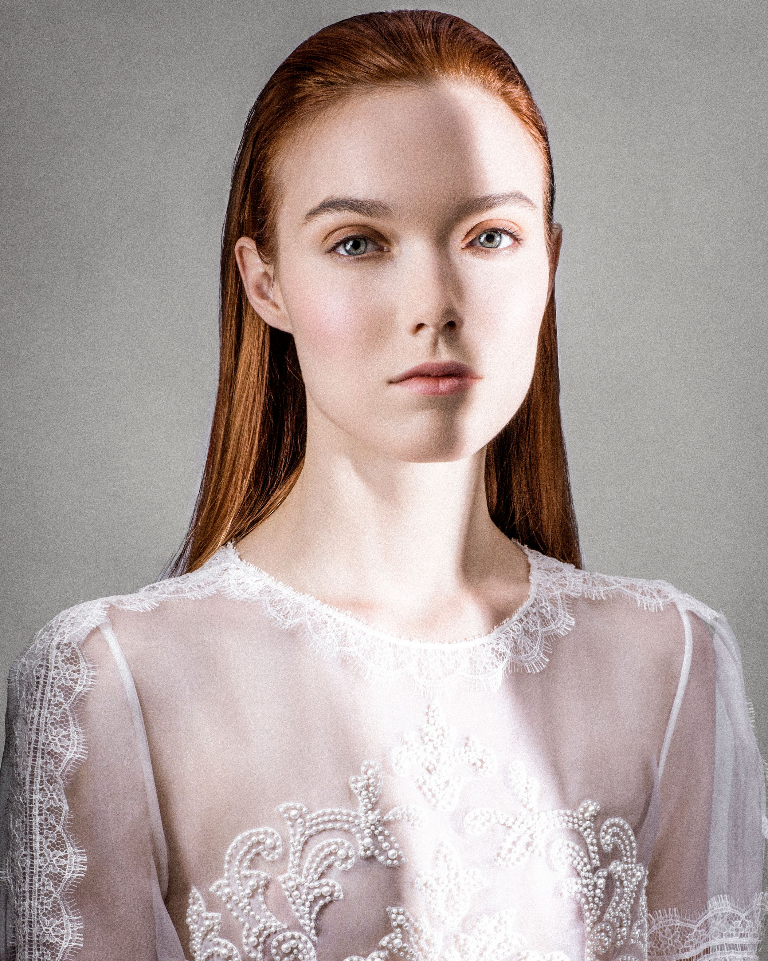 Young woman with long red hair wearing a white lace sheer top poses against a gray background.