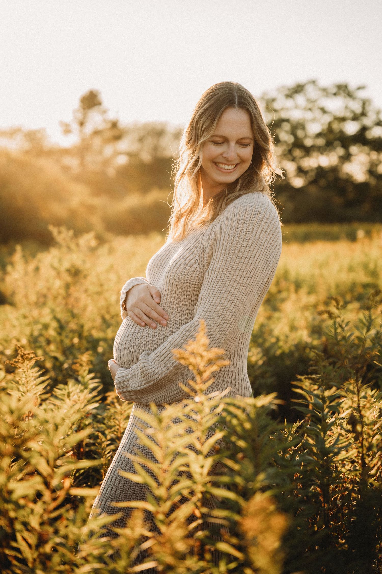 Smiling pregnant woman in beige knit dress cradling her belly in a sunlit golden field at sunset.