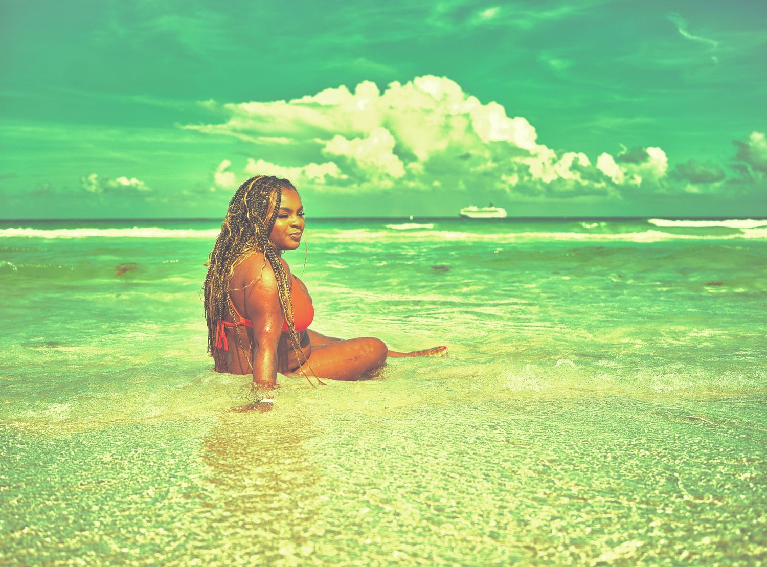 Woman sitting in shallow turquoise ocean waves on a tropical beach under a vibrant green-tinted sky with white clouds.