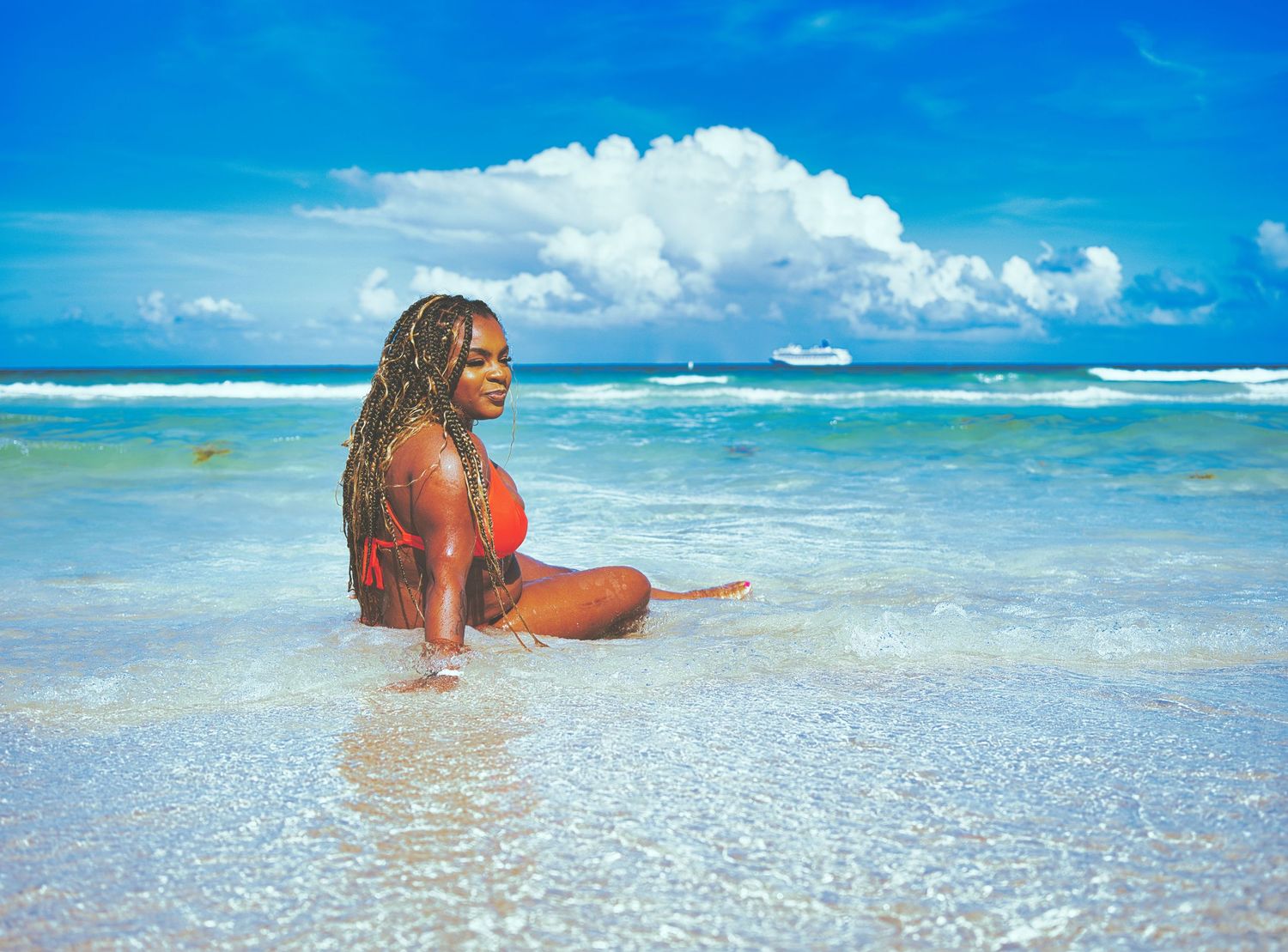 Woman in red bikini sitting in shallow turquoise ocean water on a tropical beach with a cruise ship in the distance.