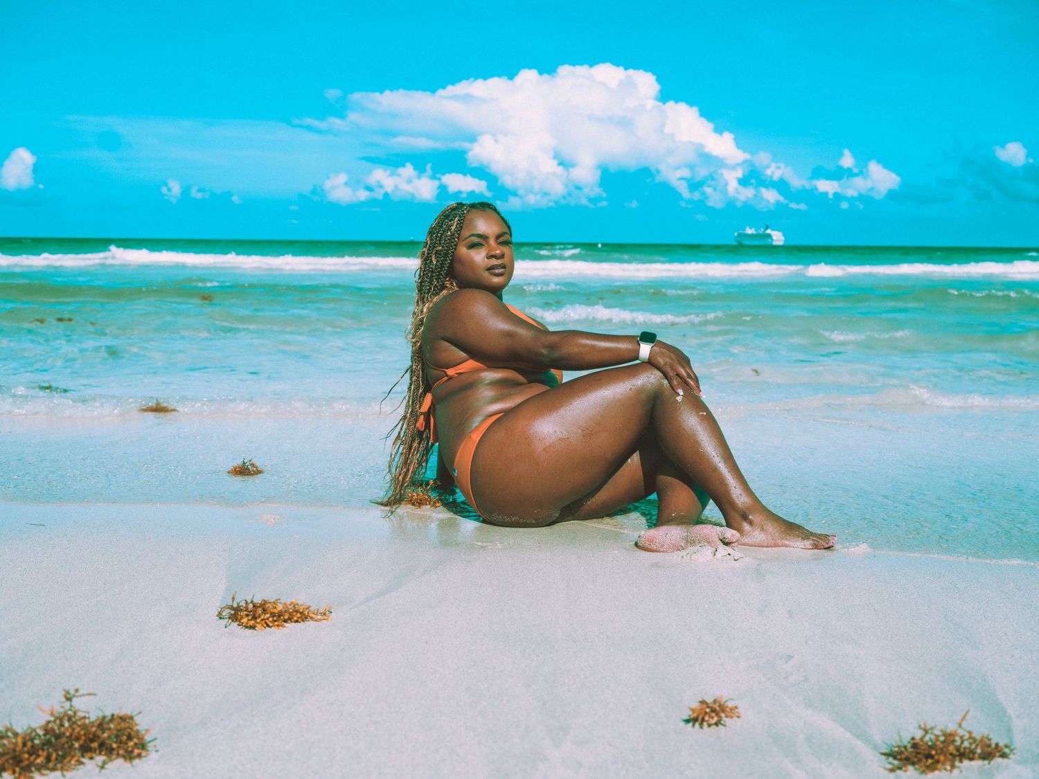 Woman in orange bikini posing on a white sand beach with turquoise ocean waters and blue sky backdrop.