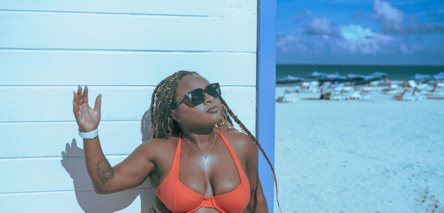 Woman in orange bikini and sunglasses posing against white wall on sunny beach with ocean in background.
