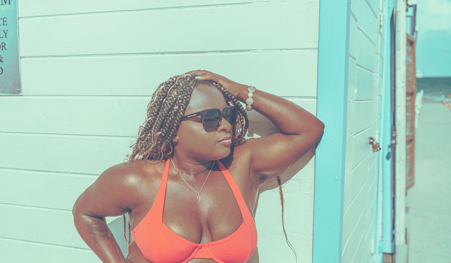 A confident Black woman in an orange bikini poses against a teal wall, wearing sunglasses and braids.