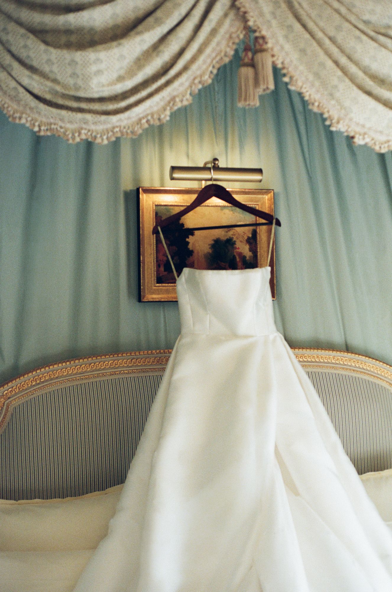 Elegant white wedding dress hanging on a wooden hanger over a gold-framed painting in a luxurious room.