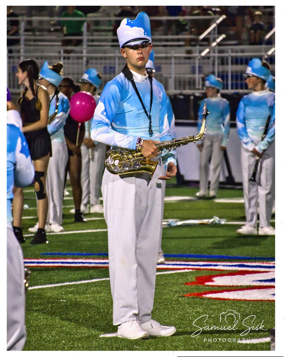 Marching band member in light blue uniform playing saxophone on a football field.