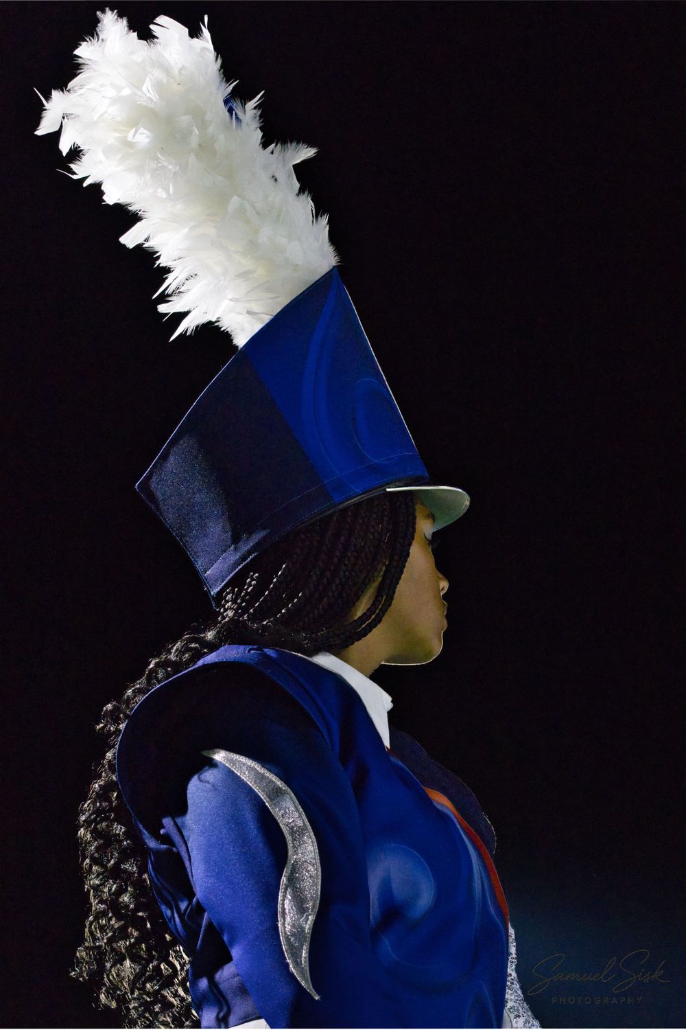 Profile view of marching band member in royal blue uniform with tall hat and white feather plume.