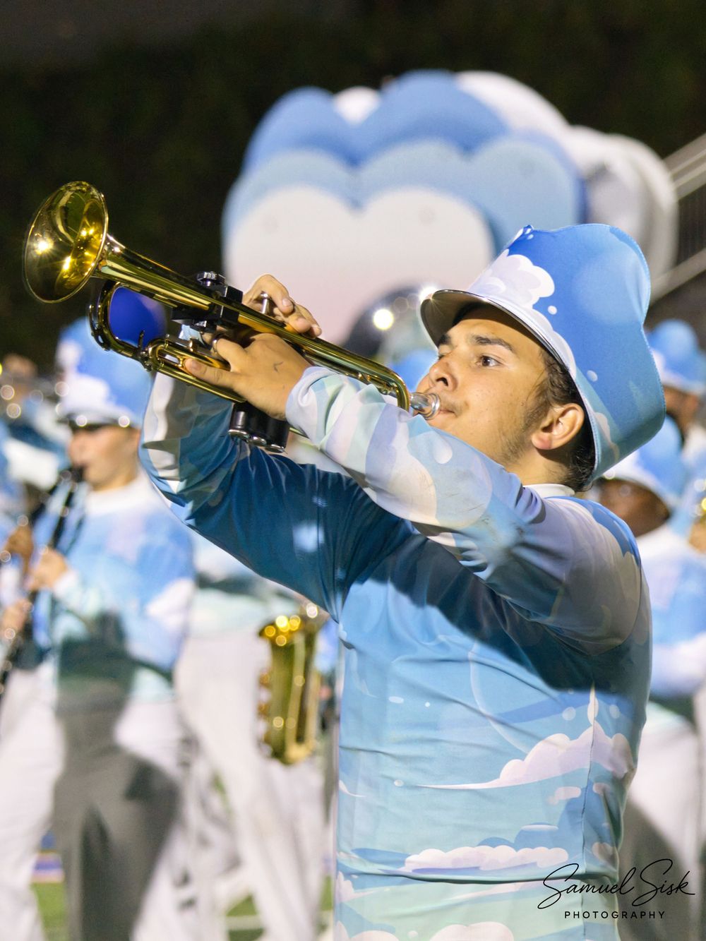 Marching band trumpet player in sky blue cloud-themed uniform performing on field at night.