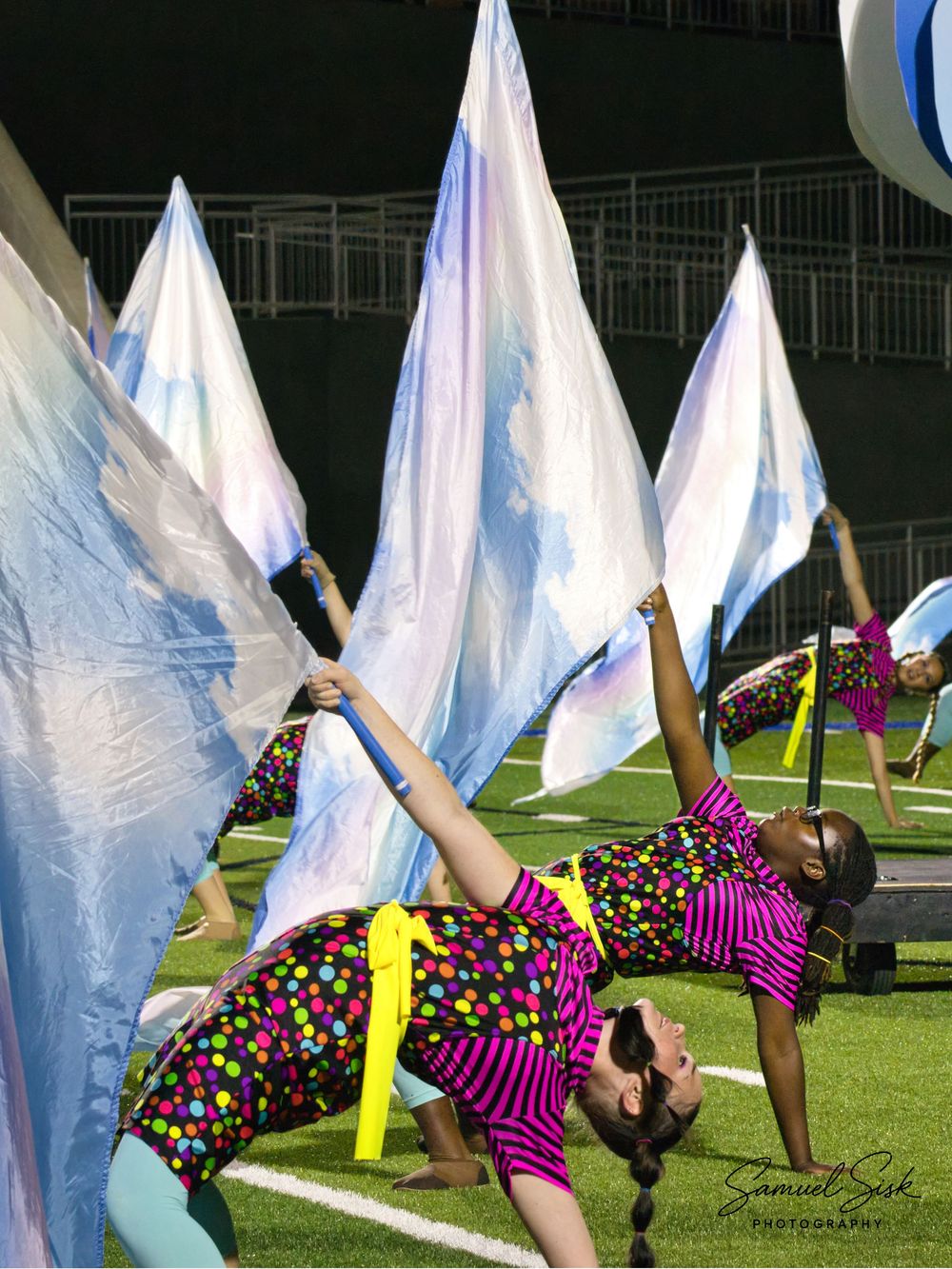 Color guard performers in colorful costumes spinning large iridescent flags on a football field at night.