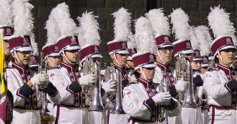 Marching band members in white and maroon uniforms playing brass instruments during a night performance.