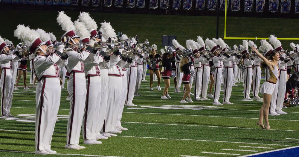 Marching band in white and maroon uniforms performing on green football field during daytime.