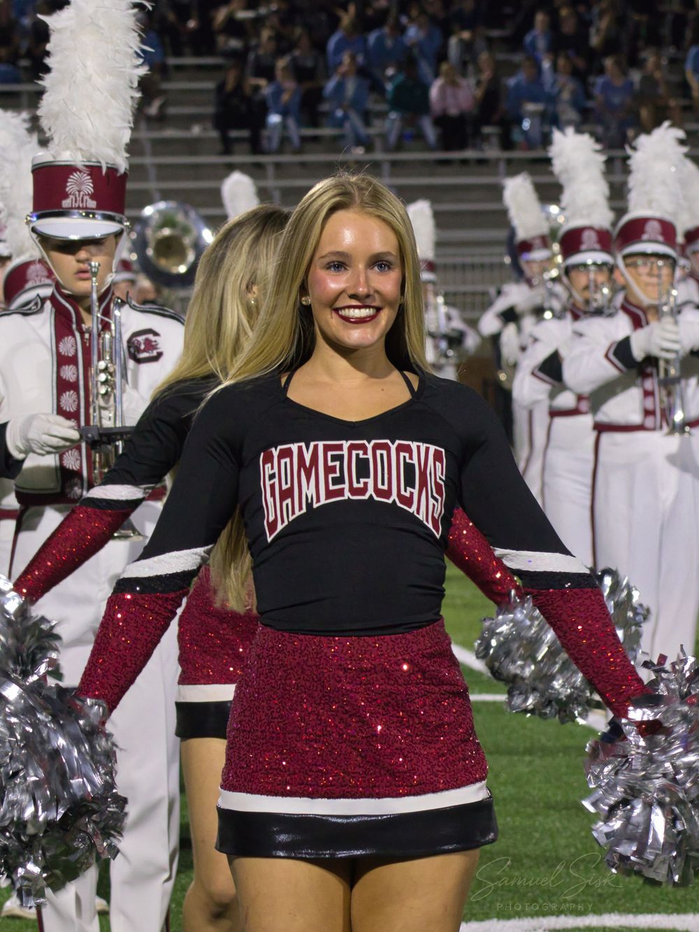 Smiling Gamecocks cheerleader in sequin skirt with marching band performing in background.