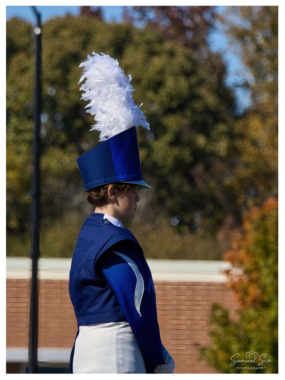 Marching band member in blue uniform with white feather-plumed shako hat outdoors in autumn.