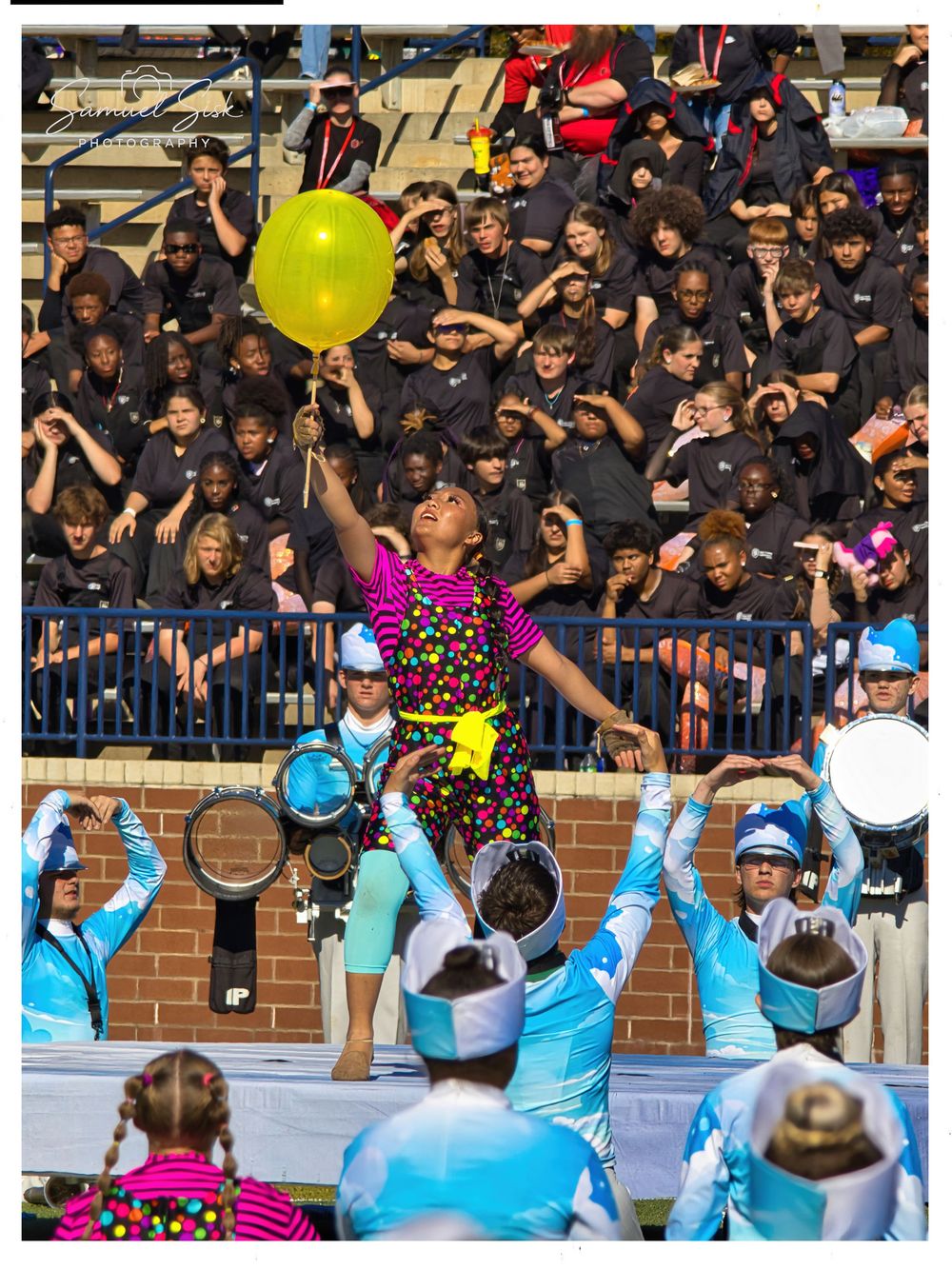 Color guard performer tossing yellow balloon above crowd of band members in black uniforms.