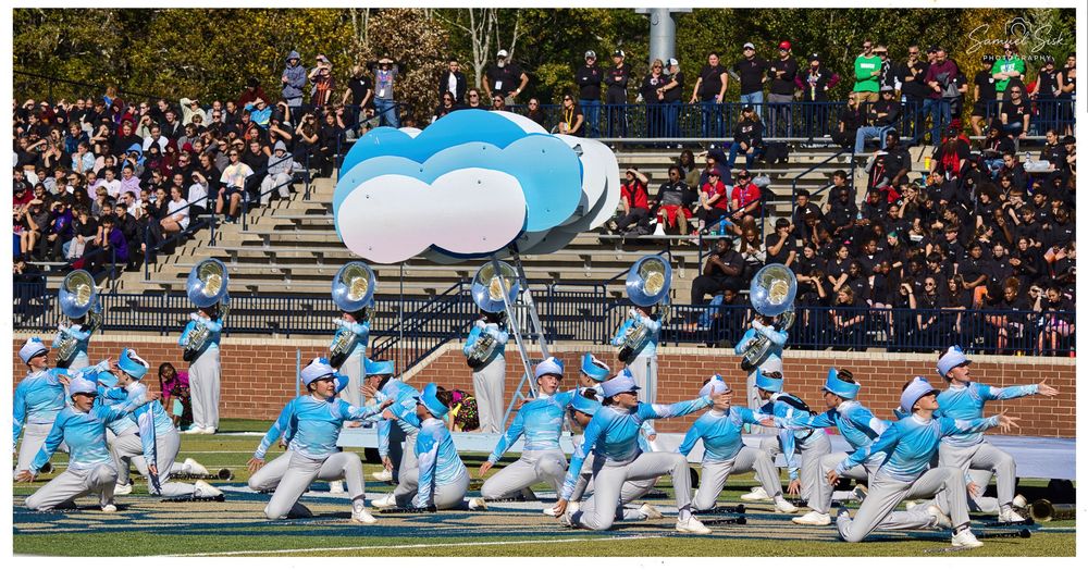 Marching band performs in blue cloud uniforms with large balloon props at an outdoor stadium competition.