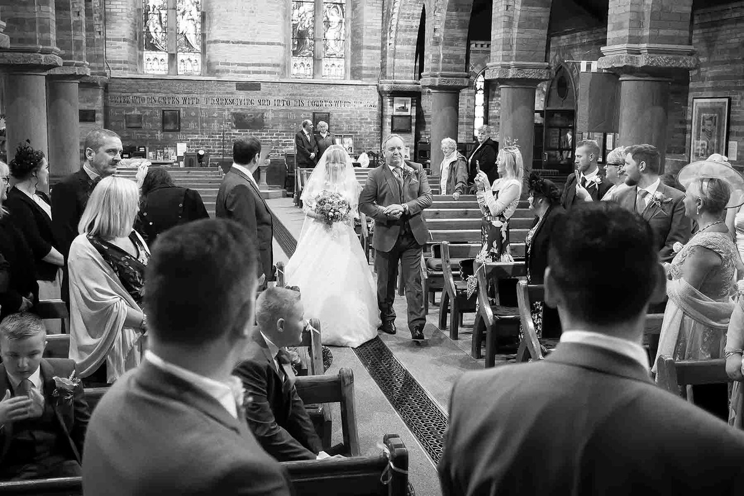 Black and white wedding photo of bride walking down church aisle with guests seated in pews during ceremony.