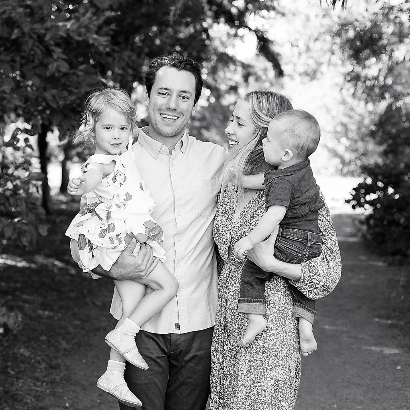 Black and white family portrait of smiling couple holding two young children outdoors on a tree-lined path.