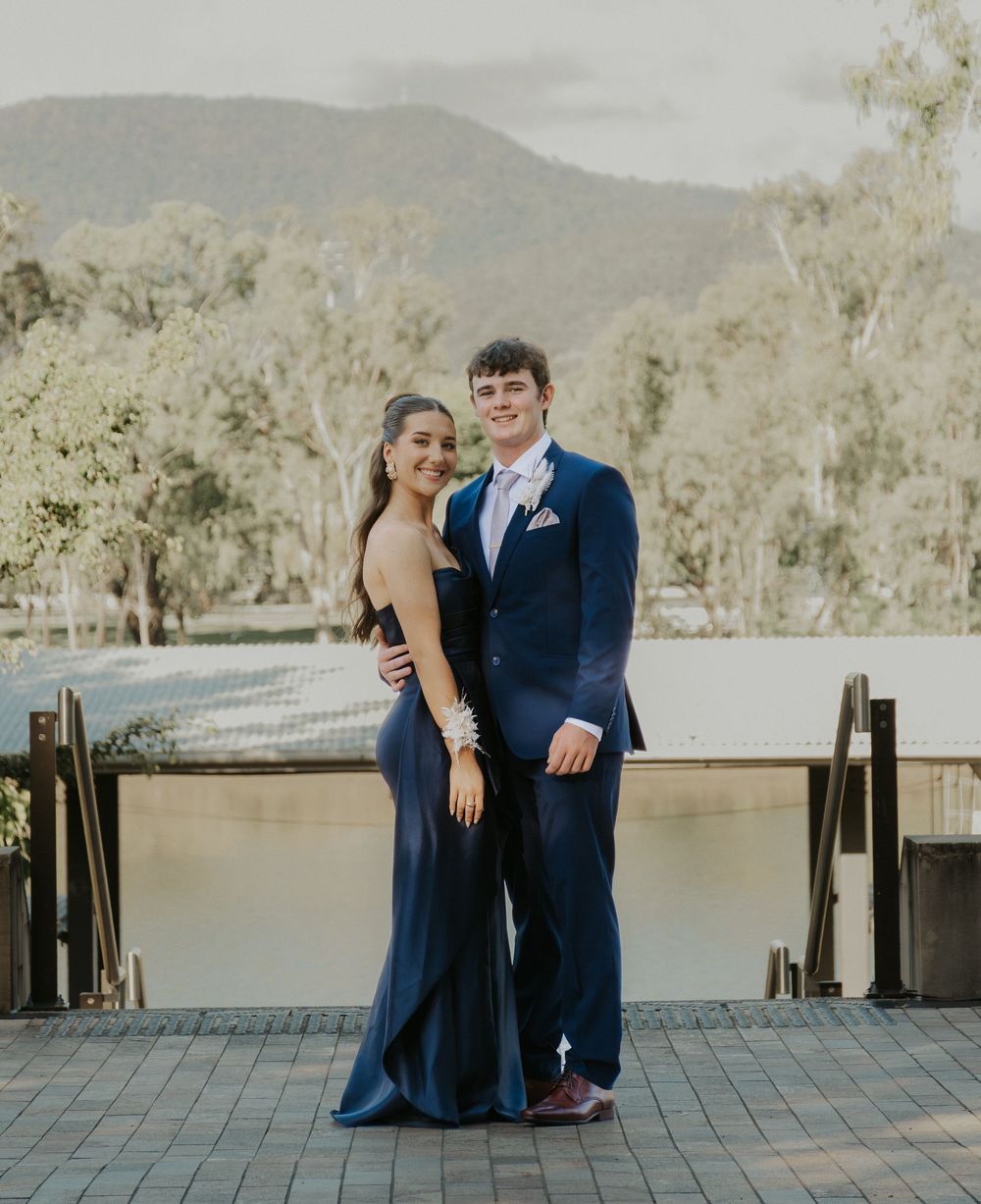 Couple in matching navy blue formal wear posing together at an outdoor prom venue with mountain views.