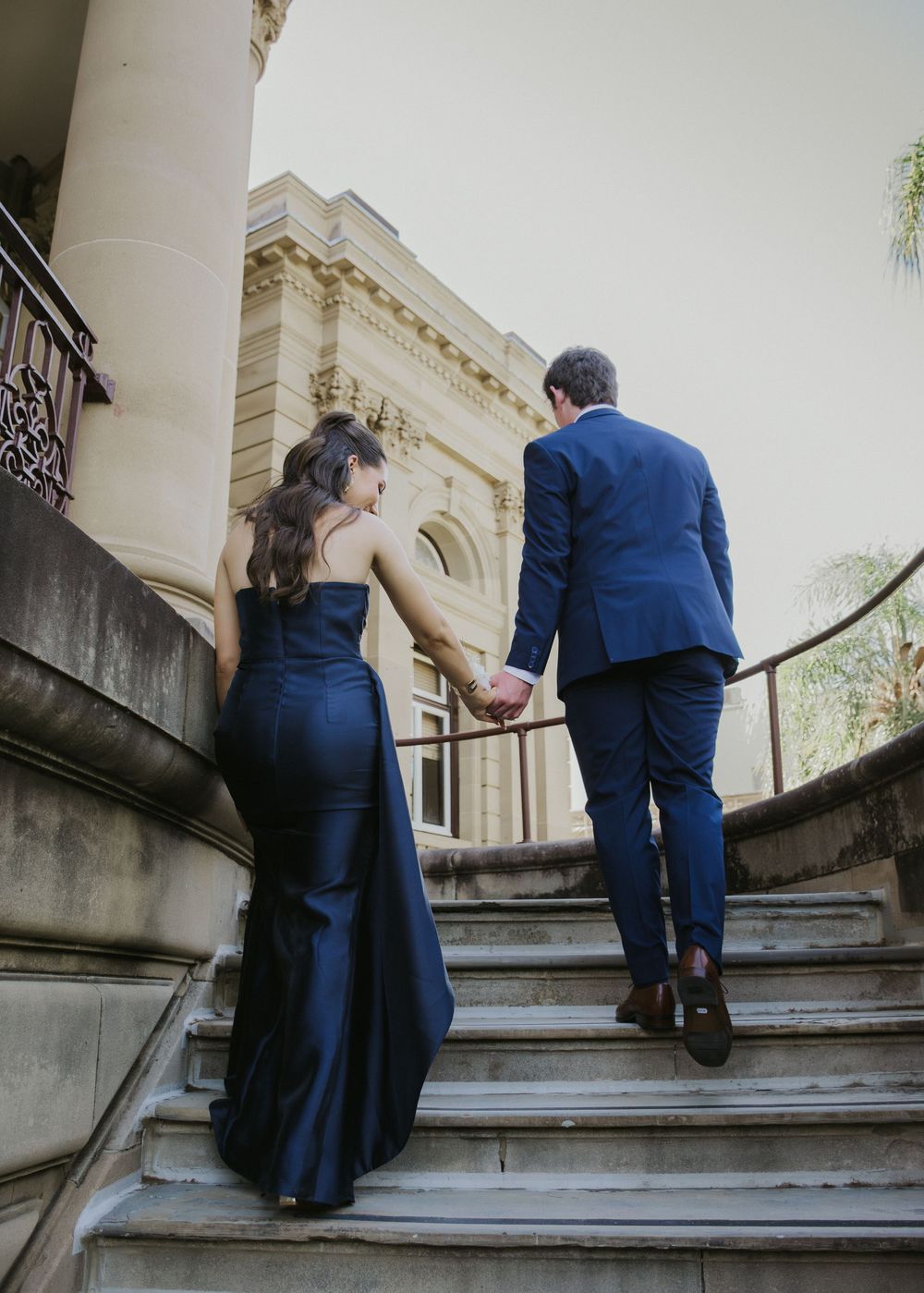 Couple in elegant navy formal wear holding hands while ascending grand stone staircase at historic venue.