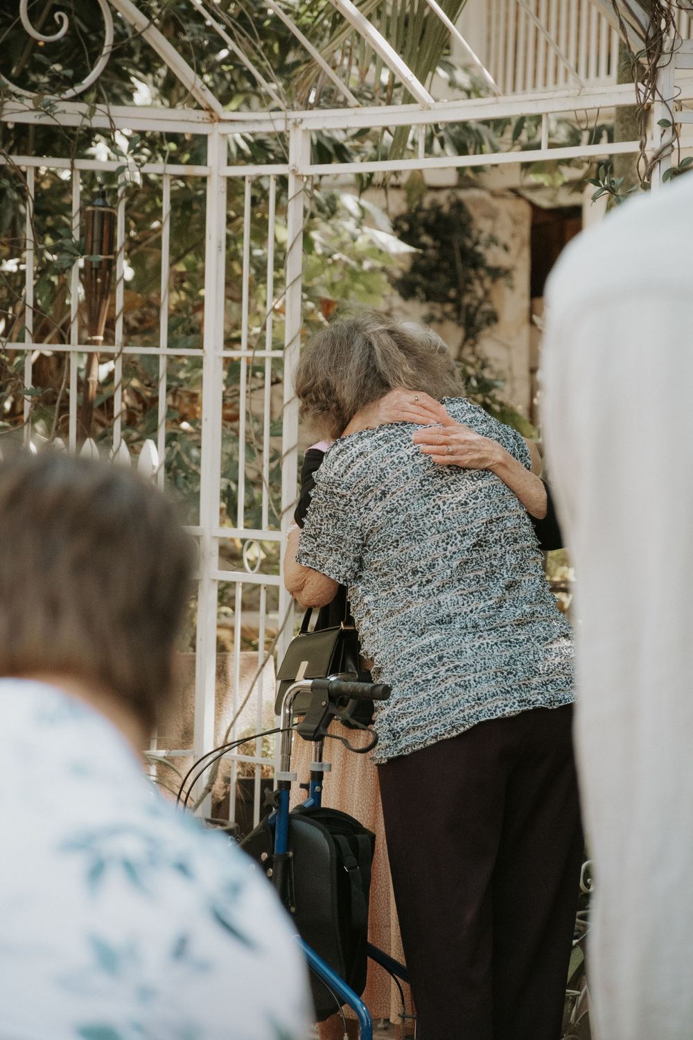 Elderly woman with walker receiving a warm hug near white garden gate.