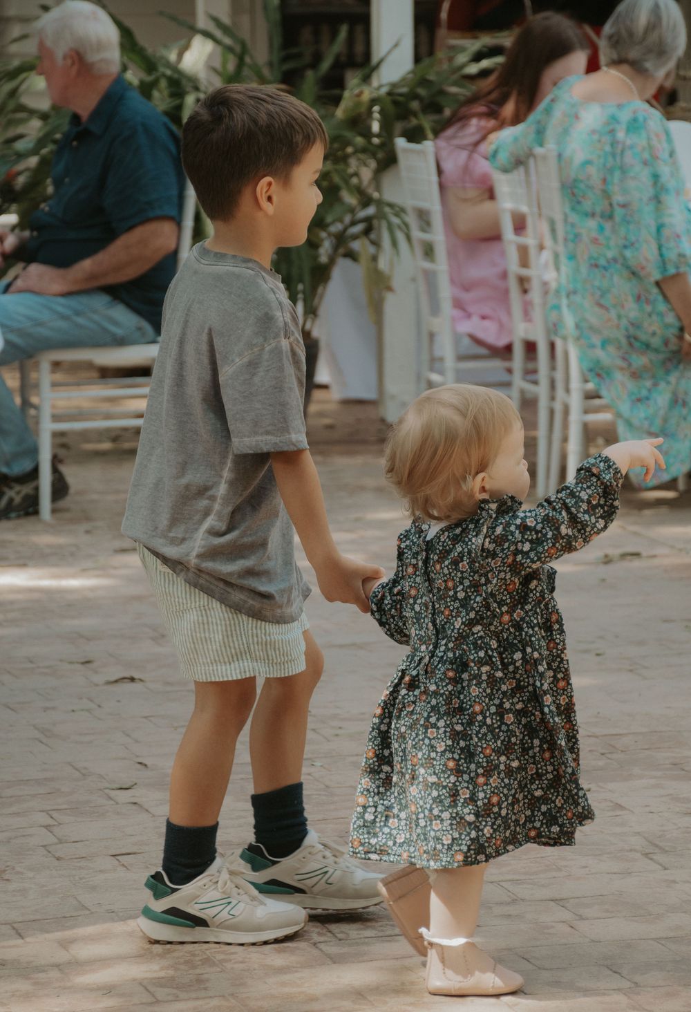 Young boy holding hands with toddler girl in floral dress at outdoor event.