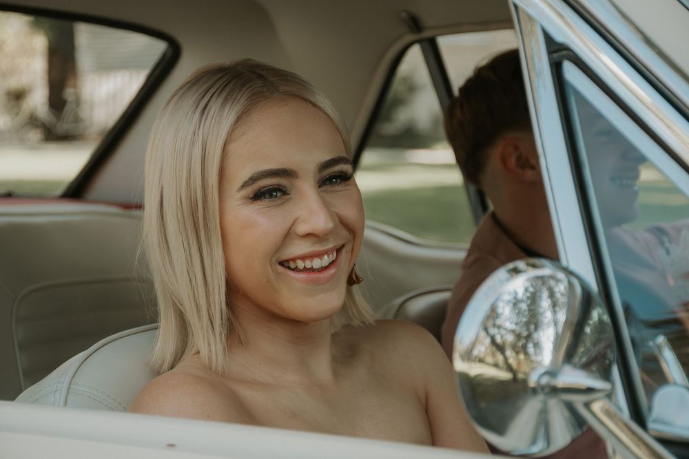 Smiling blonde woman in strapless dress sitting in vintage car interior looking over her shoulder.