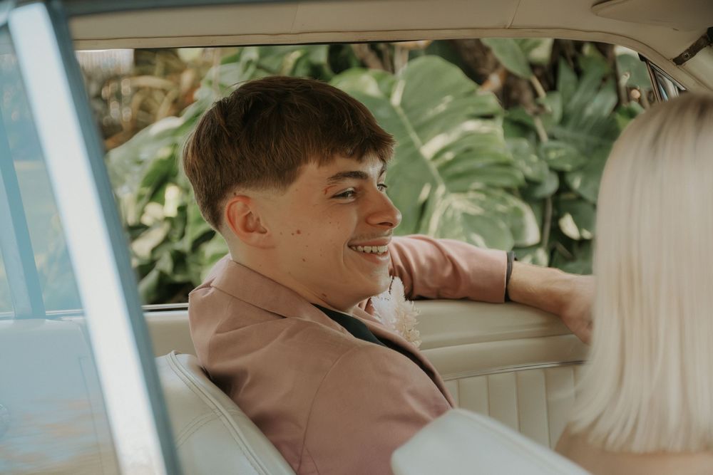 Smiling young man in pink suit relaxing in vintage car backseat surrounded by tropical foliage.