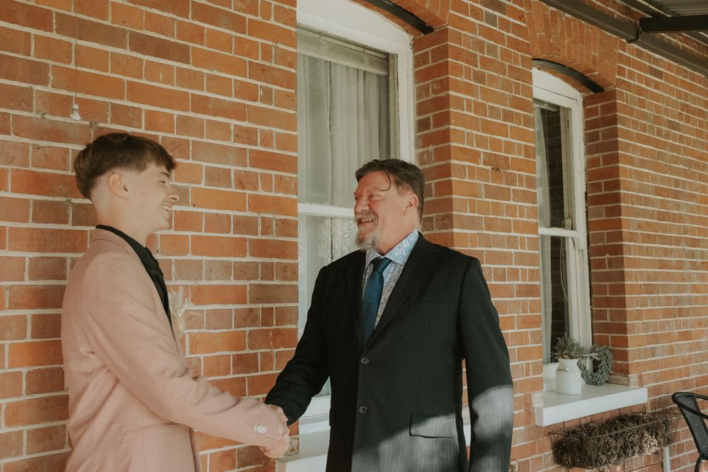 Two men in formal suits shaking hands outside a red brick building with white windows.
