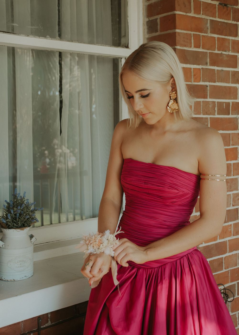 Blonde woman in strapless pink formal gown adjusting wrist corsage near vintage window display.