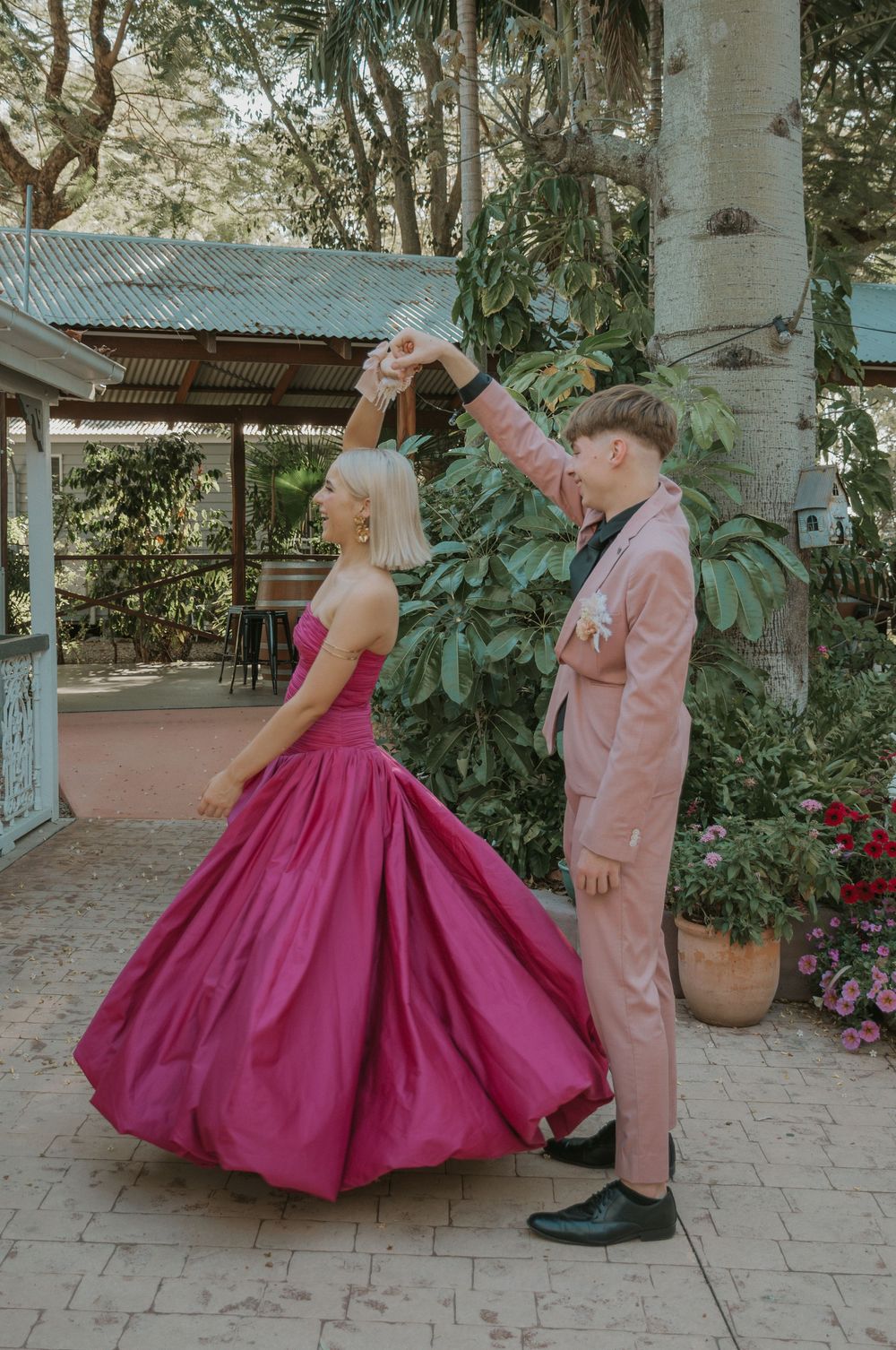 Couple dancing outdoors, woman in voluminous pink ball gown being twirled by partner in pink suit.