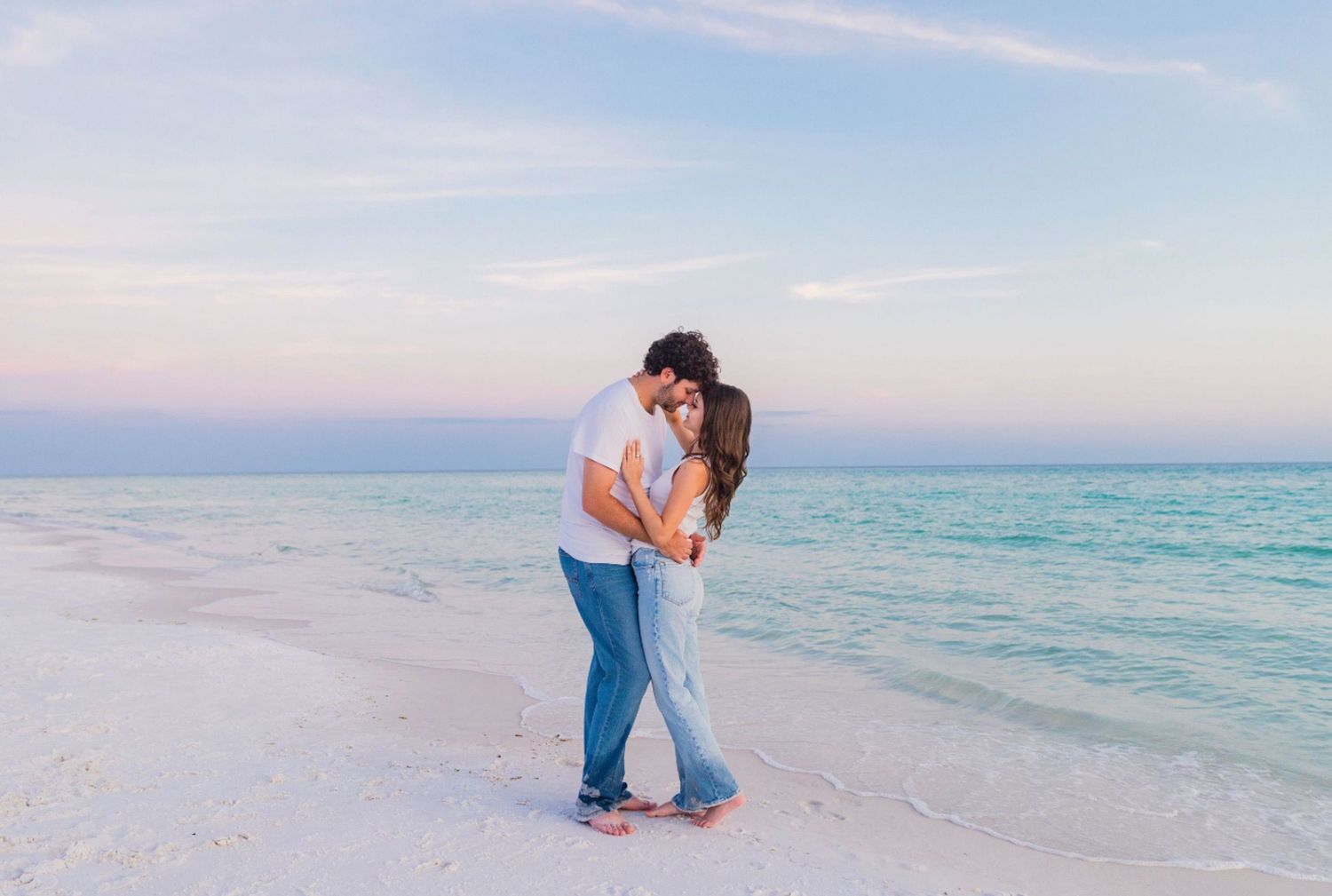 Romantic couple embracing on a white sand beach at sunset with turquoise ocean waters in the background.
