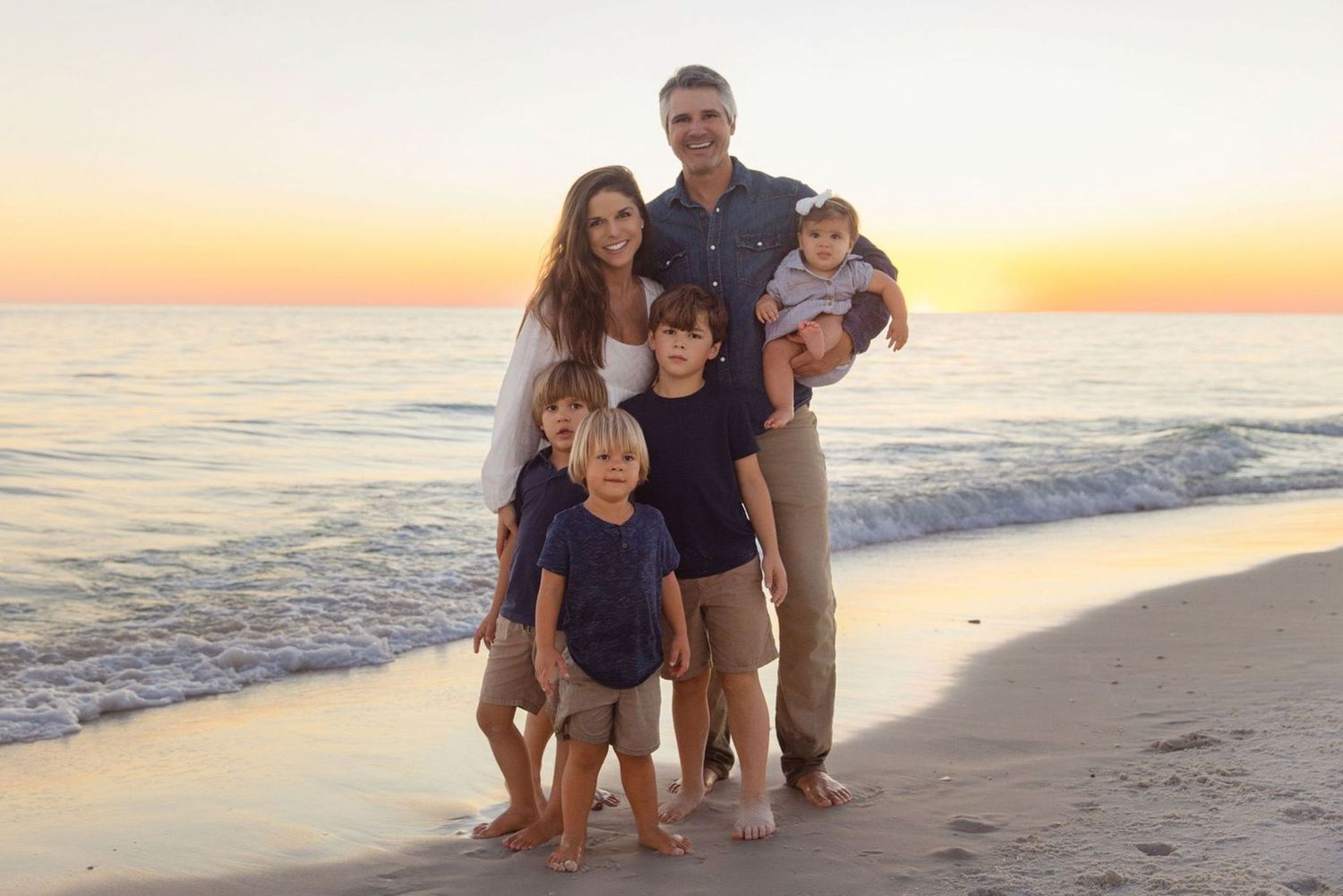 Family of six poses on a sandy beach at sunset, with calm ocean waves and a colorful sky in the background.