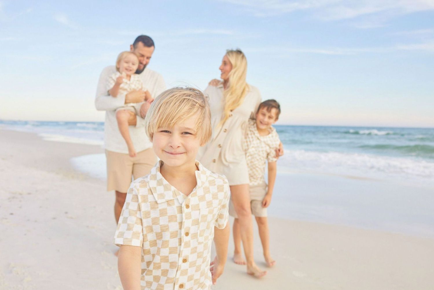 A happy young boy smiles on a sunny beach while his family poses together near the ocean shoreline behind him.
