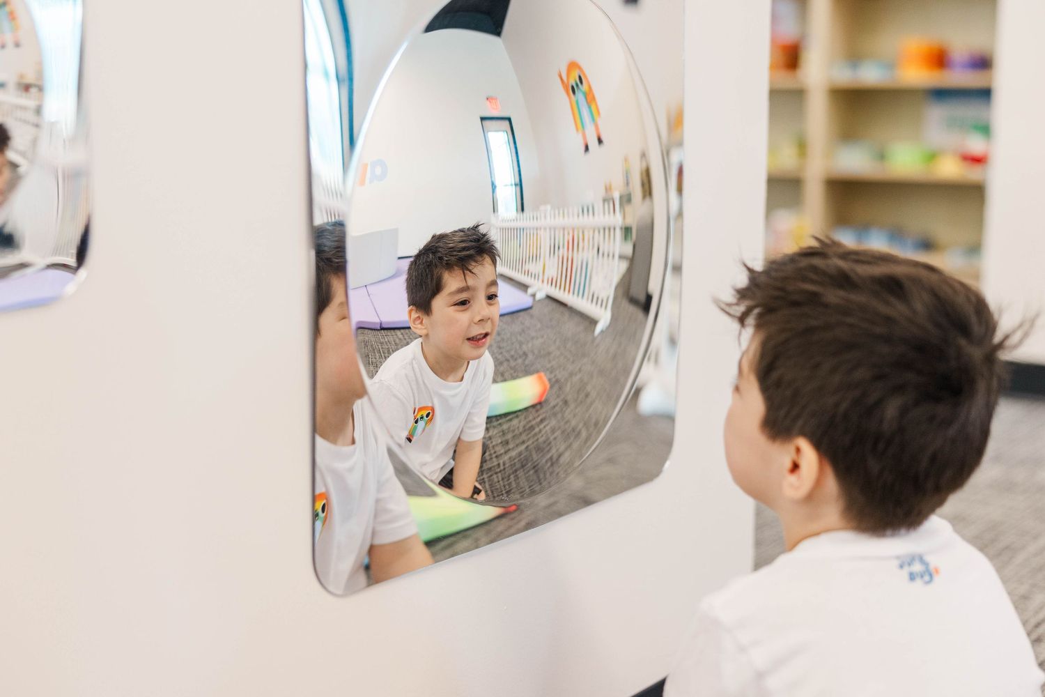 Young boy looking at his reflection in a rounded mirror in a bright classroom.
