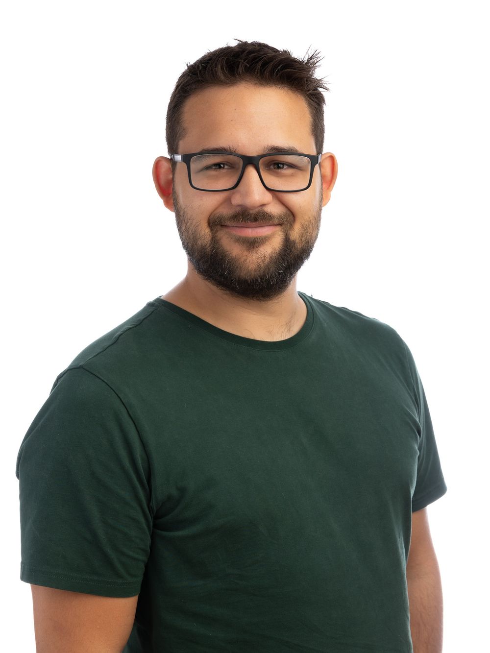 A smiling young man with glasses and a beard wearing a dark green t-shirt against a white background.