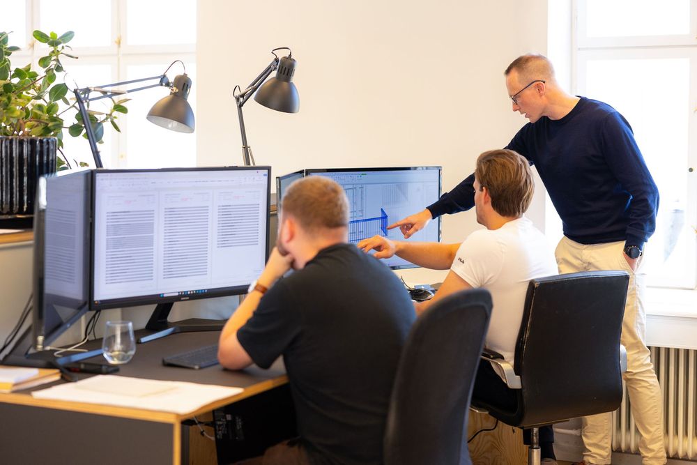 Three office professionals collaborating at dual monitors, reviewing documents in a bright modern workspace.