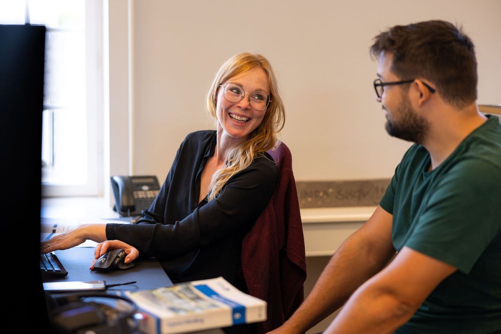 Smiling blonde woman and bearded man collaborating at a computer workstation in an office.