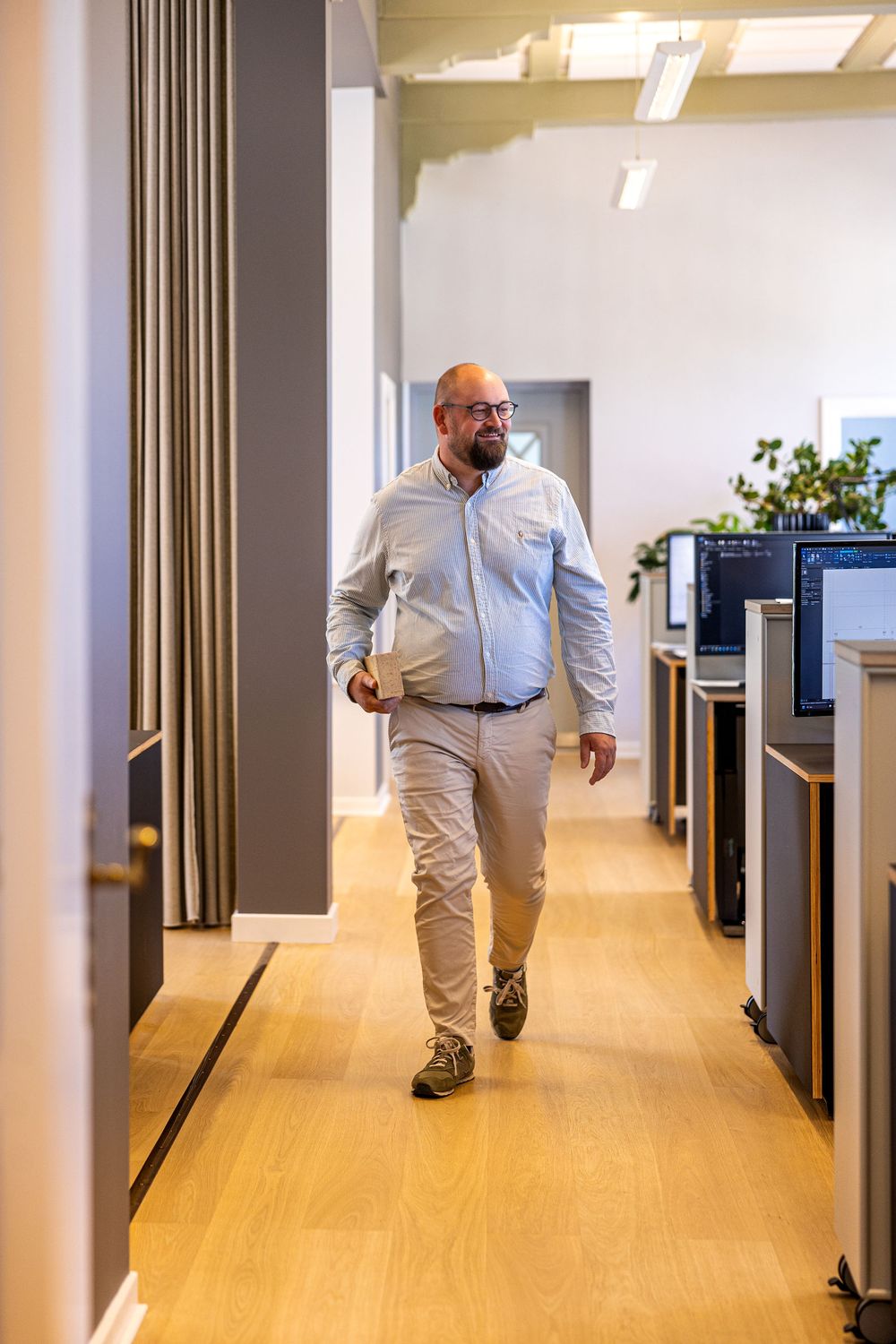 Bald man with glasses walks through modern office hallway carrying laptop, wearing light blue shirt and khaki pants.
