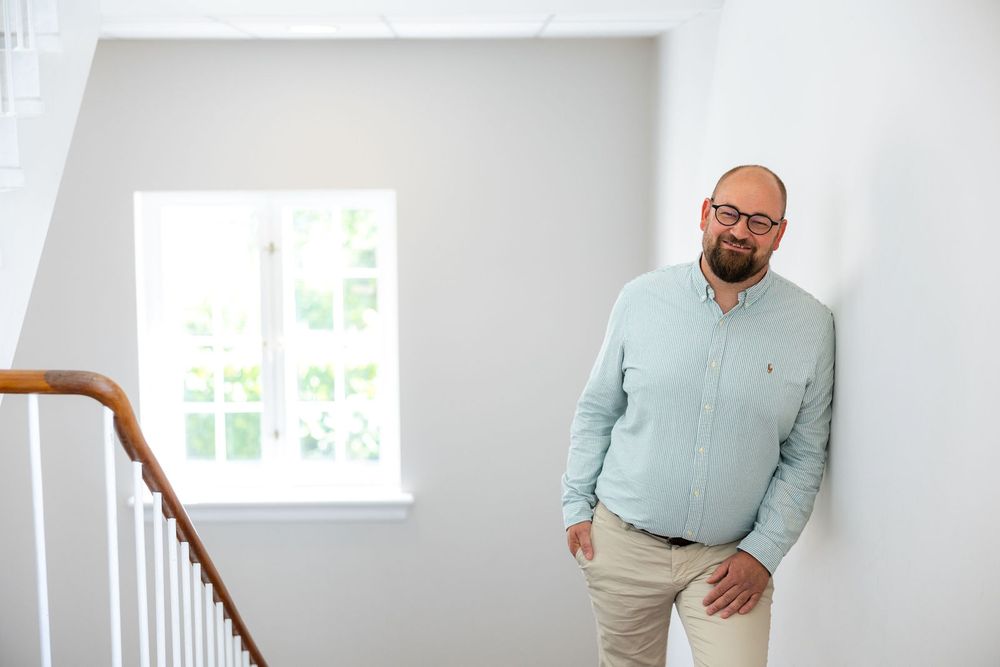 Bearded man in light blue sweater leaning against white wall near staircase in bright modern interior.