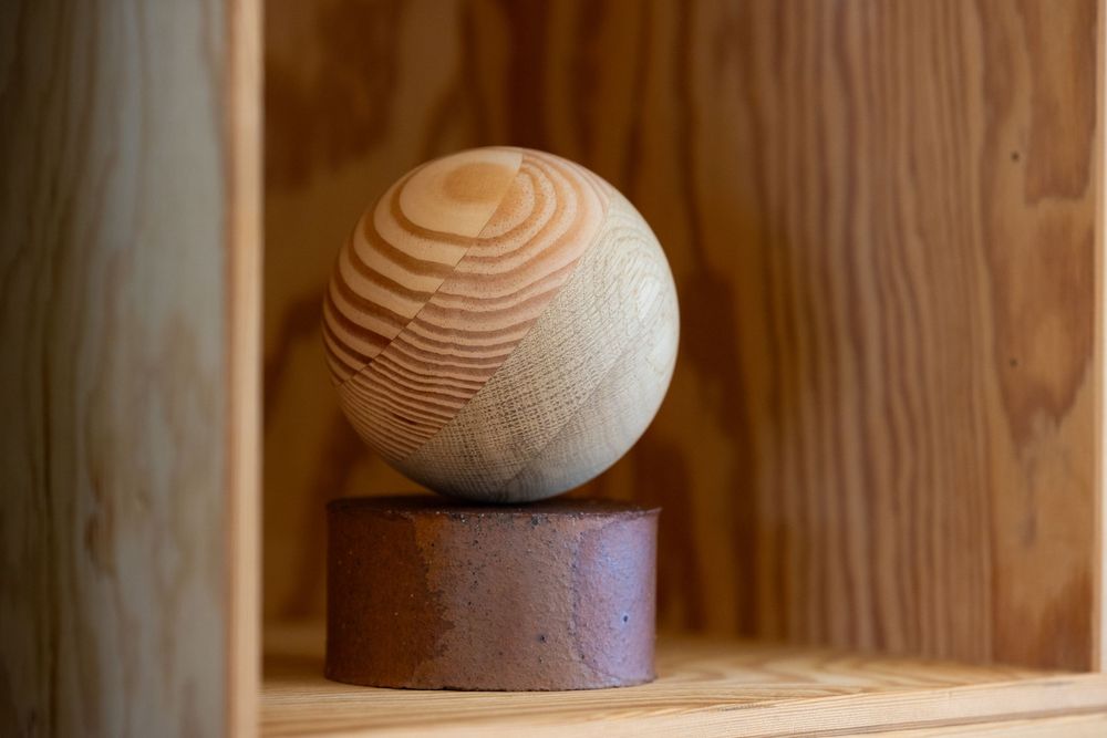 A handcrafted wooden sphere with natural grain patterns resting on a rustic brown cylindrical base on a wooden shelf.
