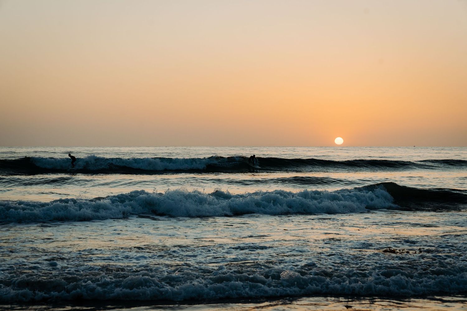 Surfers riding ocean waves at sunset with golden sky and sun on the horizon at a scenic beach.