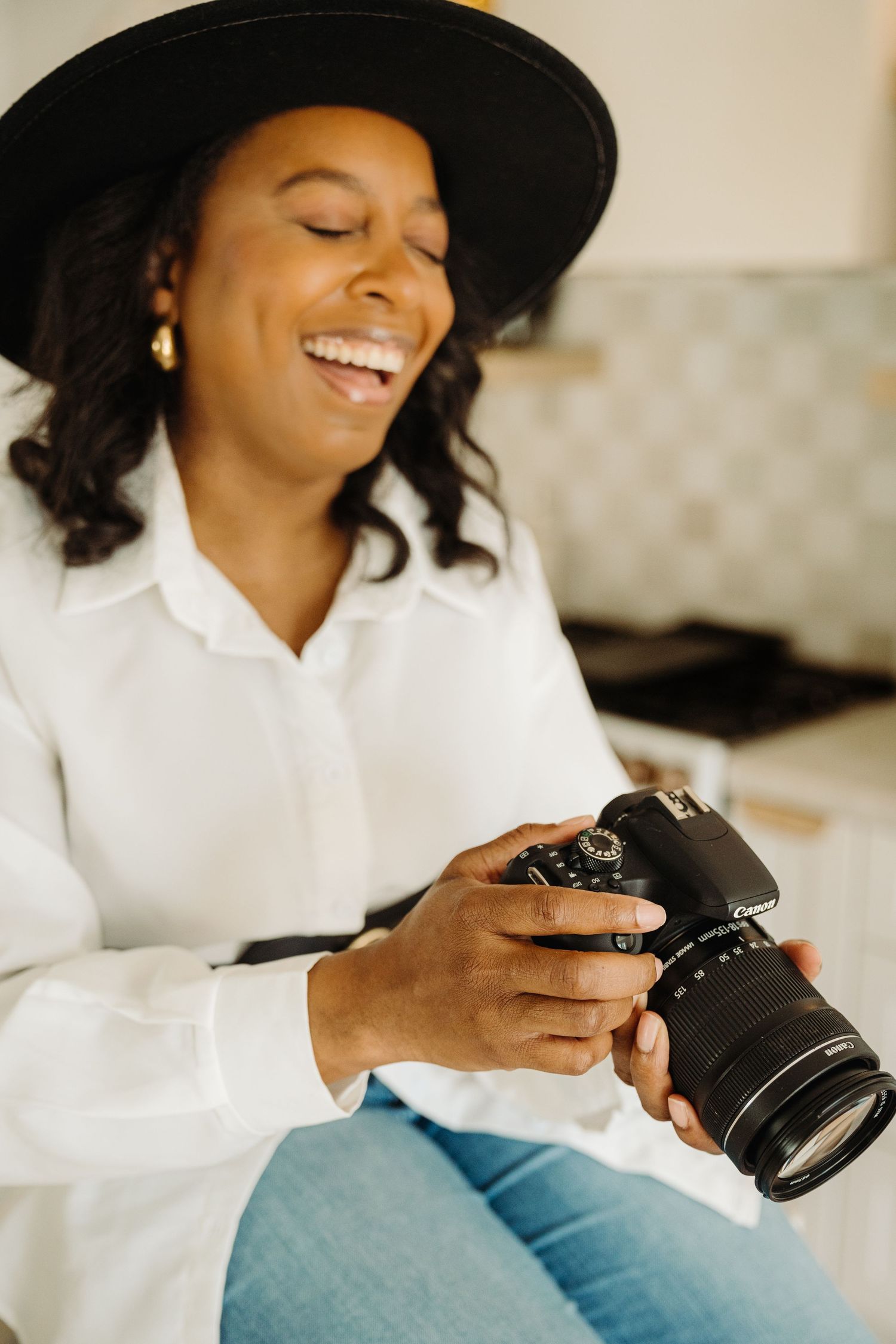 A smiling woman in a white shirt and black hat holds a Canon camera while sitting in a bright kitchen.
